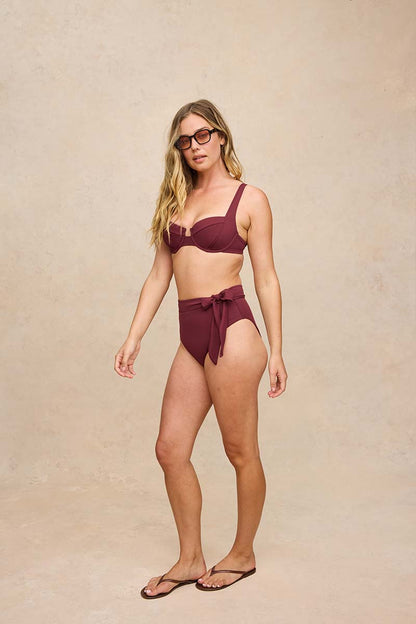 A woman modeling a two-piece high-waisted bikini in a deep burgundy color, paired with dark sunglasses and flip-flops, standing against a textured beige studio backdrop.