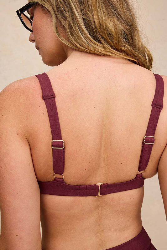 Rear view of a woman wearing a burgundy adjustable sports bra with gold-tone metal buckles and straps, set against a neutral beige background.
