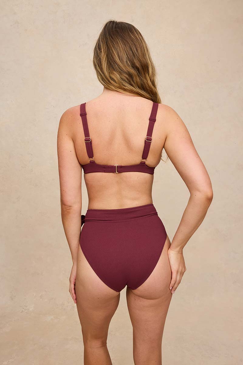 A woman shown from the back wearing a two-piece swimsuit in a deep burgundy color, featuring a high-waisted bottom and a strappy top with adjustable buckles, set against a textured beige background.