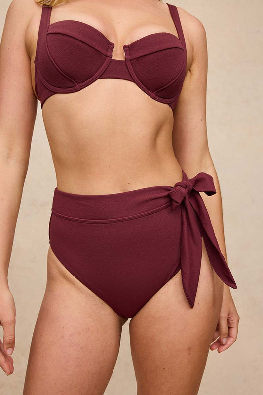 A woman wearing a two-piece bikini in deep burgundy, featuring a structured underwire bra top and high-waisted side-tie bottoms, shown against a neutral beige background.