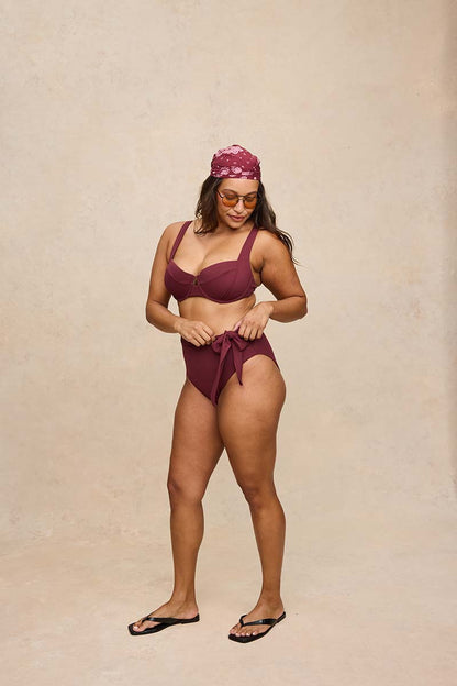 A woman modeling a two-piece swimsuit consisting of a deep burgundy bikini top and matching high-waisted bikini bottoms with a side tie, paired with a matching burgundy headscarf and black flip-flops, against a textured beige studio backdrop.