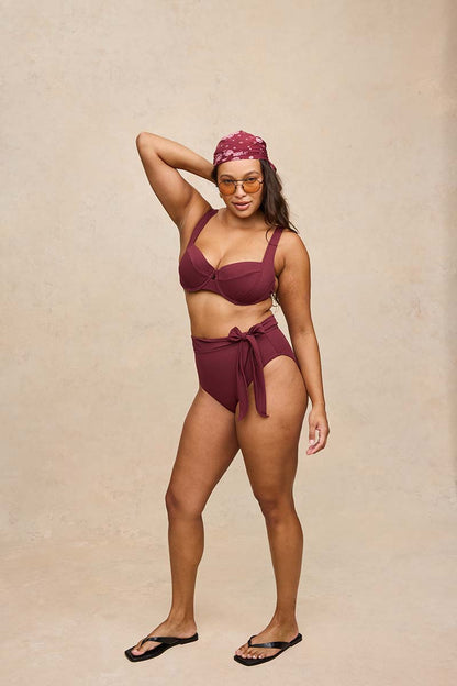 A woman modeling a two-piece burgundy swim set with a matching headscarf and yellow-tinted sunglasses, standing against a textured beige studio backdrop.