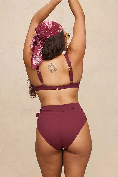 A woman shown from the back wearing a two-piece bikini in a deep burgundy color, paired with a matching burgundy paisley headscarf. She is standing against a neutral beige textured wall.