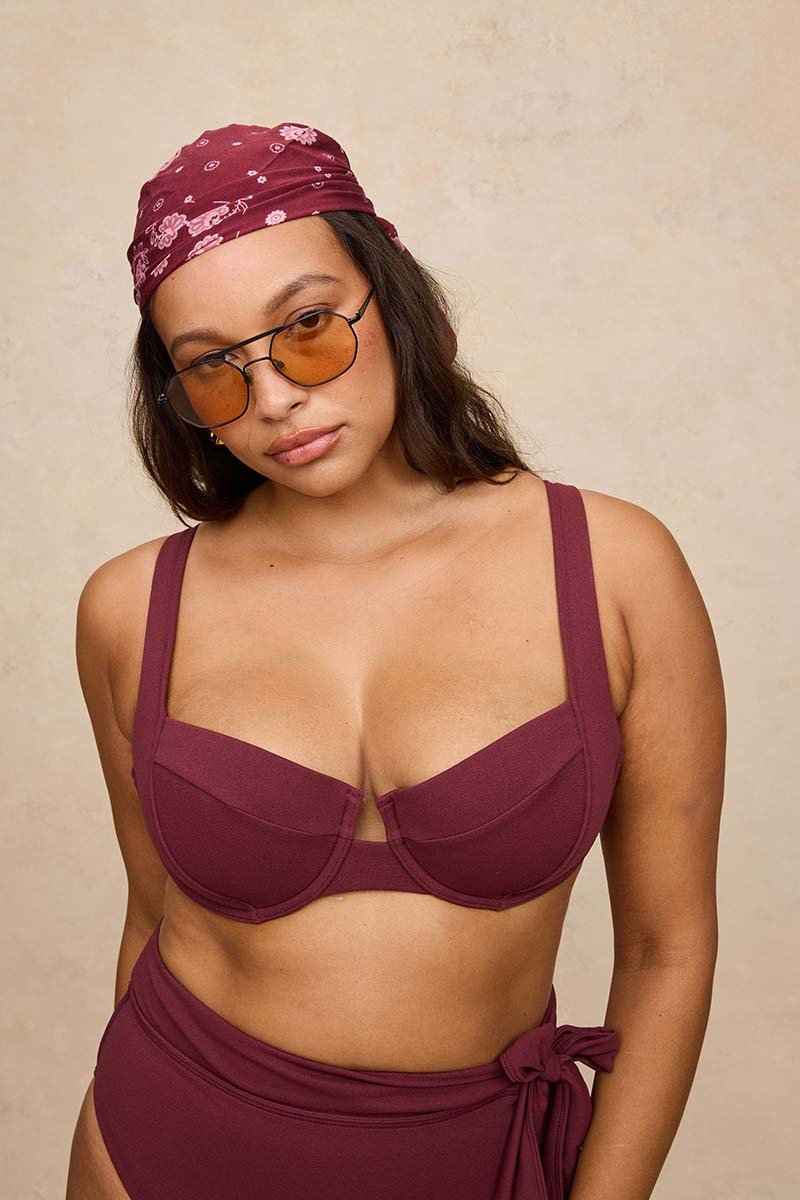 A woman modeling a matching burgundy two-piece swimwear set, featuring a bra top with wide straps and high-waisted bottoms with a side tie, paired with a matching patterned headscarf and tortoiseshell sunglasses, against a textured beige background.