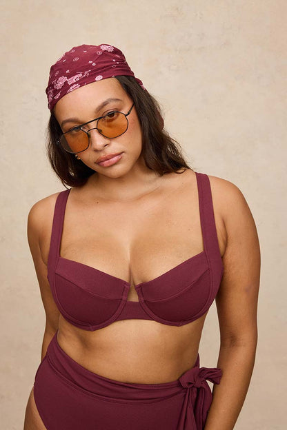 A woman modeling a matching burgundy two-piece swimwear set, featuring a bra top with wide straps and high-waisted bottoms with a side tie, paired with a matching patterned headscarf and tortoiseshell sunglasses, against a textured beige background.