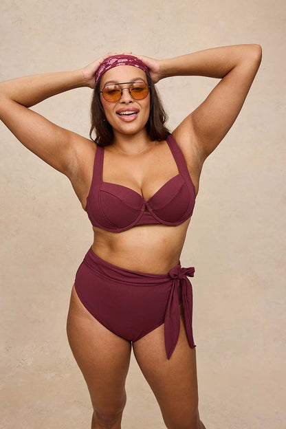 A woman modeling a two-piece bikini in a deep burgundy/maroon color, featuring a bandeau-style top and high-waisted bottoms with a side tie. She is wearing orange-tinted sunglasses and a matching burgundy headband against a neutral beige textured background.