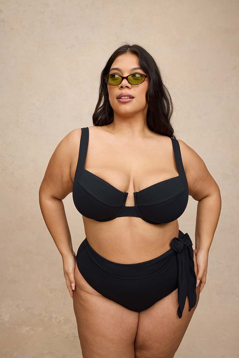 A plus-size model wearing a black two-piece bikini with a side-tie detail, styled with olive green sunglasses, standing against a textured beige background.