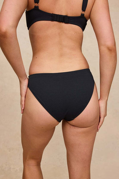 Rear view of a model wearing a black bikini bottom and matching black bikini top against a warm beige background.