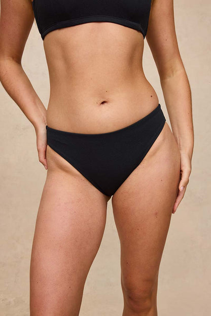 A model wearing a pair of black high-waisted bikini bottoms, shown from the chest down against a neutral beige background.
