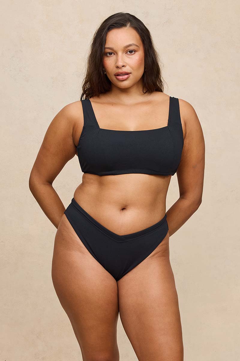 A woman modeling a black two-piece bikini set against a textured beige studio background.