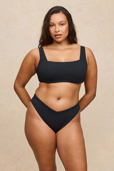 A woman modeling a black two-piece bikini set against a textured beige studio background.