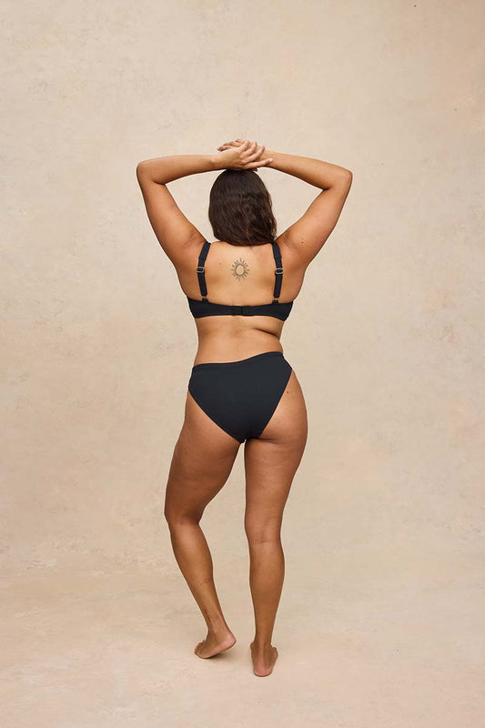 A woman is shown from behind wearing a black two-piece bikini, standing against a textured beige studio backdrop.