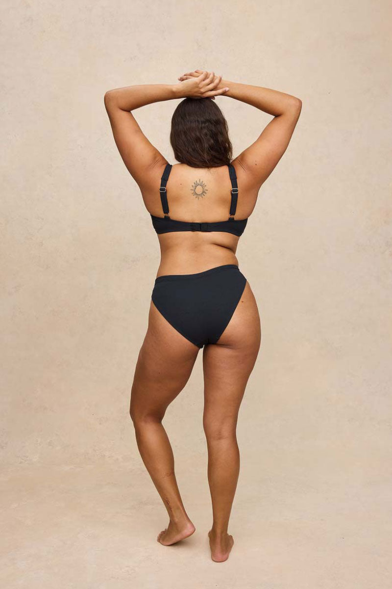 A woman is shown from behind wearing a black two-piece bikini set against a textured beige studio backdrop.