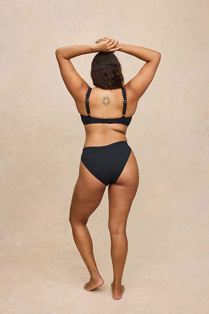 A woman is shown from behind wearing a black two-piece bikini set against a textured beige studio backdrop.