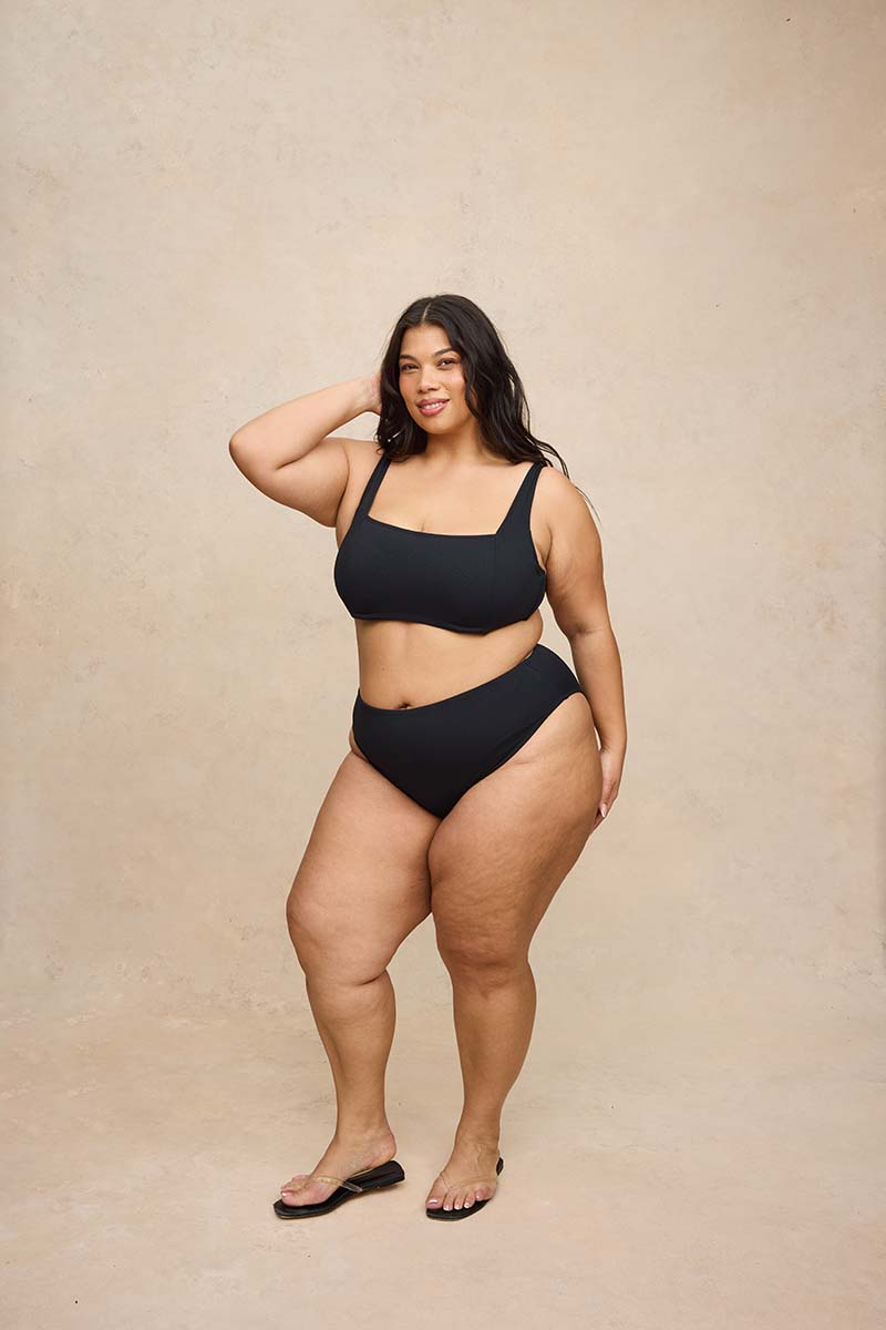 A plus-size woman modeling a black two-piece bikini set against a textured beige studio backdrop.
