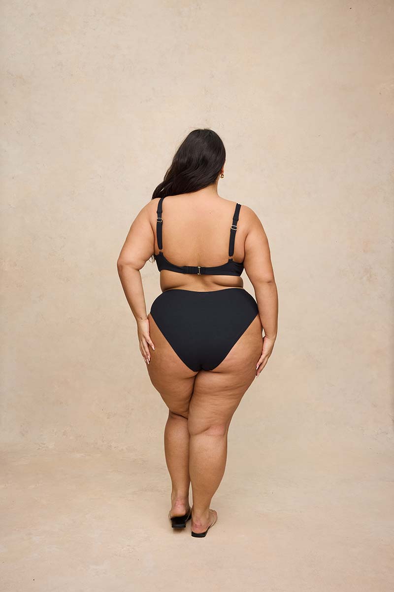 A plus-size woman is shown from behind wearing a black two-piece bikini and black flat sandals, standing against a textured beige studio backdrop.