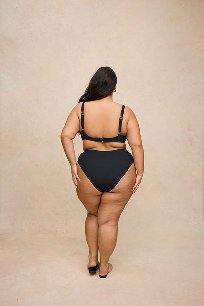 A plus-size woman is shown from behind wearing a black two-piece bikini and black flat sandals, standing against a textured beige studio backdrop.