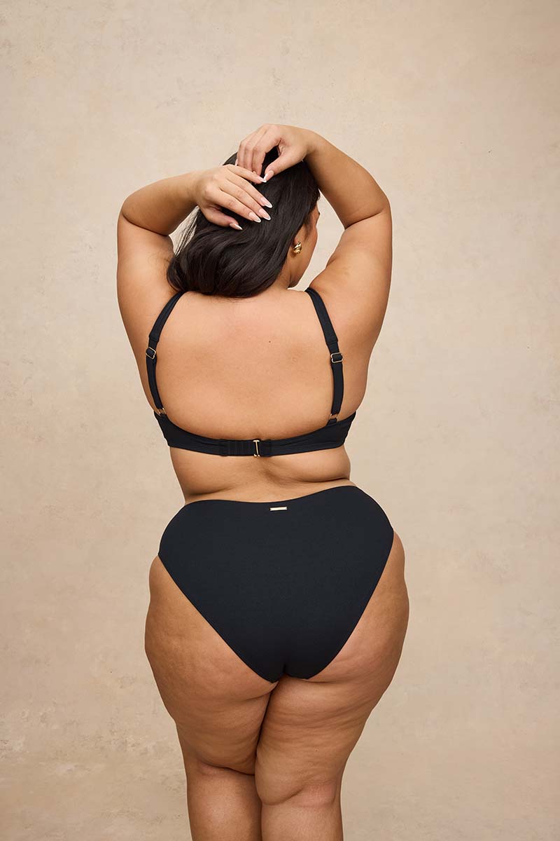 A woman shown from behind wearing a black two-piece bikini set against a textured beige studio background.