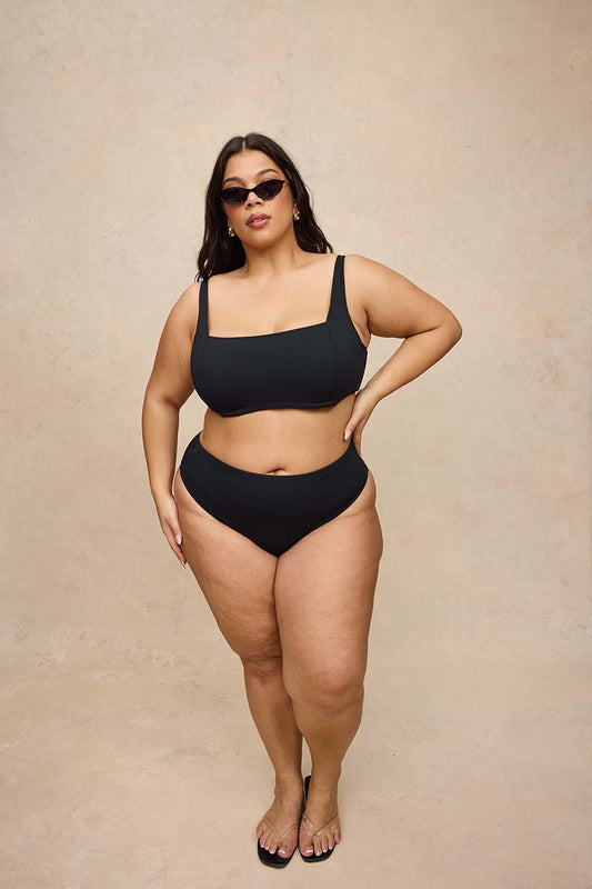 A plus-size model wearing a solid black two-piece bikini swimsuit with a square-neck top and high-waisted bottoms, styled with black sunglasses and black sandals, standing against a textured beige studio backdrop.