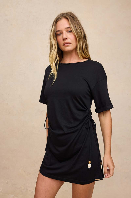 A woman modeling a black short-sleeve wrap-style mini dress with drawstring details and small decorative beads near the hem, posed against a textured beige studio backdrop.