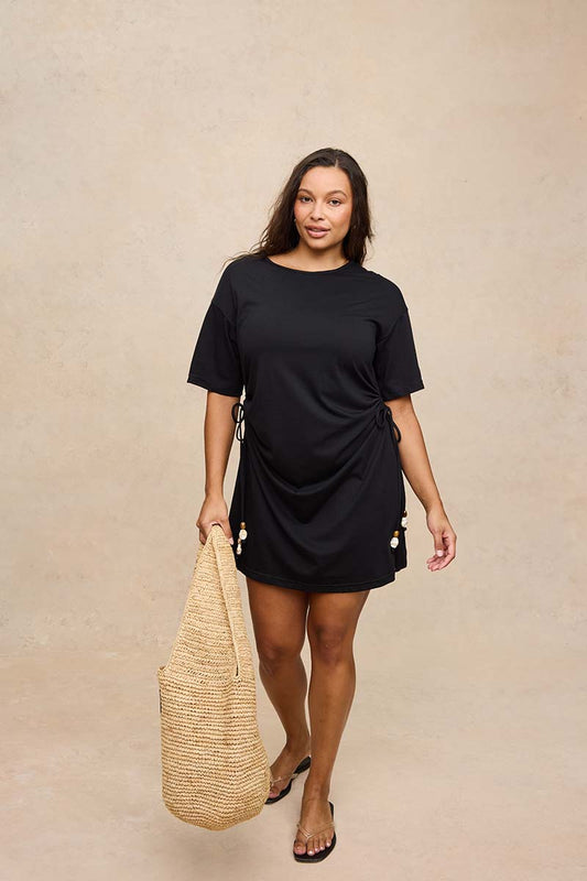 A woman modeling a black short-sleeve mini dress with side tie details and pearl accents, holding a large woven straw tote bag against a textured beige studio backdrop.
