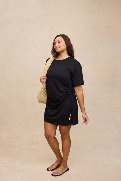 A woman modeling a black short-sleeve maternity dress with a side twist detail, carrying a woven straw shoulder bag against a textured beige studio backdrop.