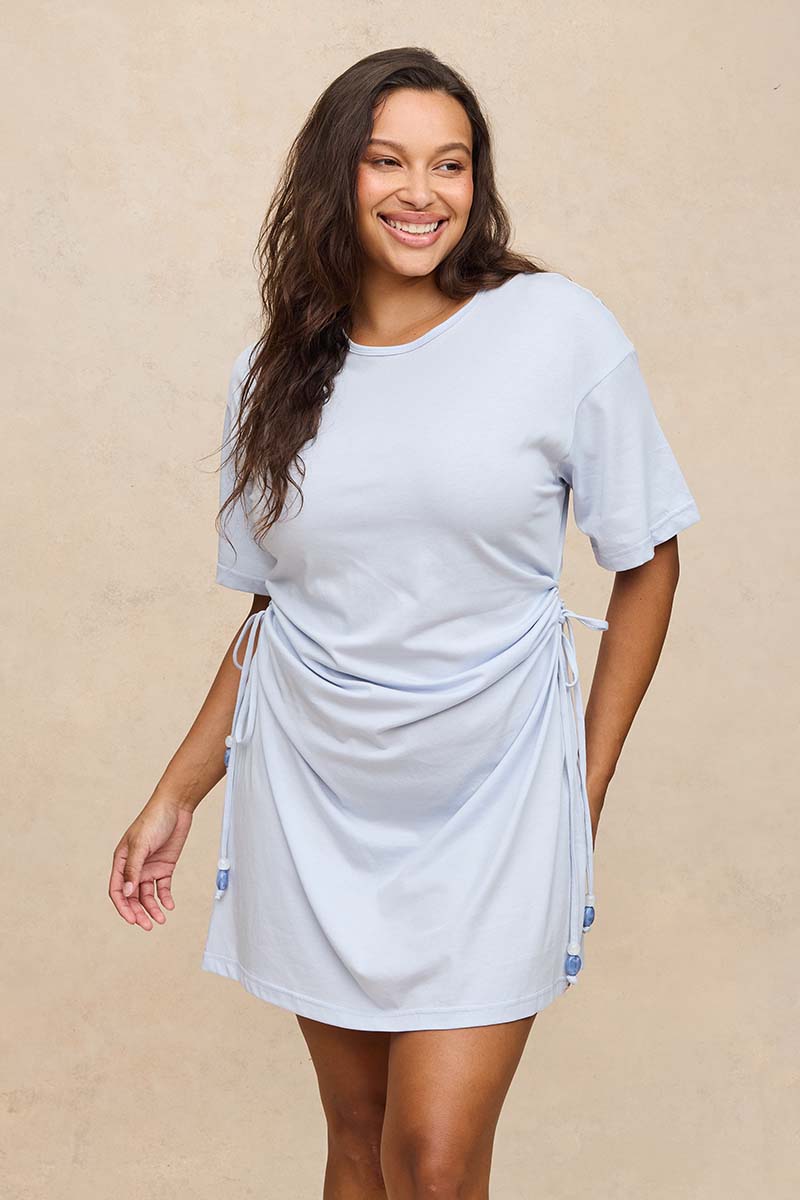 A woman modeling a light blue short-sleeve t-shirt dress with a side tie detail, set against a neutral beige textured background.