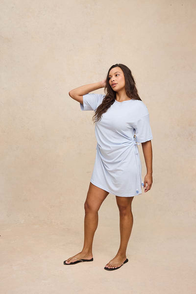 A woman modeling a light blue short-sleeve wrap-style mini dress with side tie details, standing against a textured beige studio backdrop.