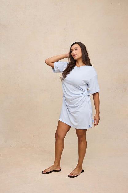 A woman modeling a light blue short-sleeve wrap-style mini dress with side tie details, standing against a textured beige studio backdrop.