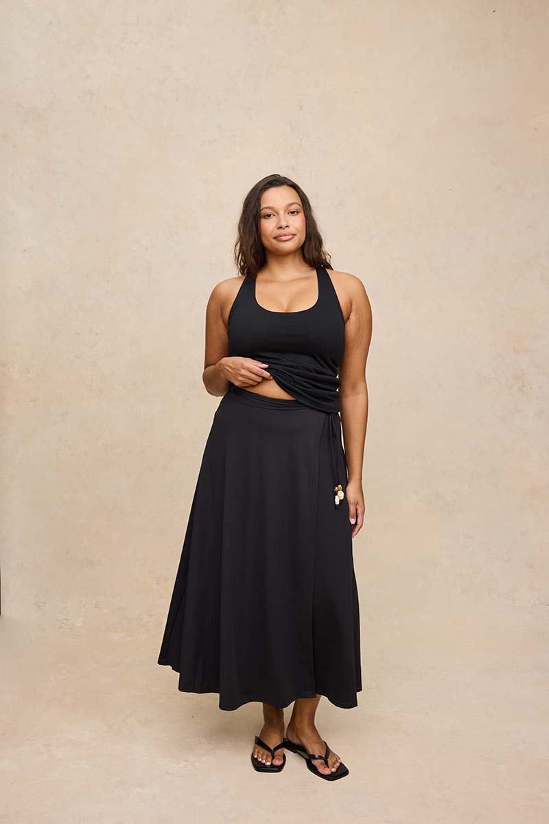 A woman modeling a black two-piece set consisting of a halter crop top and a matching high-waisted midi skirt, standing against a textured beige studio backdrop.