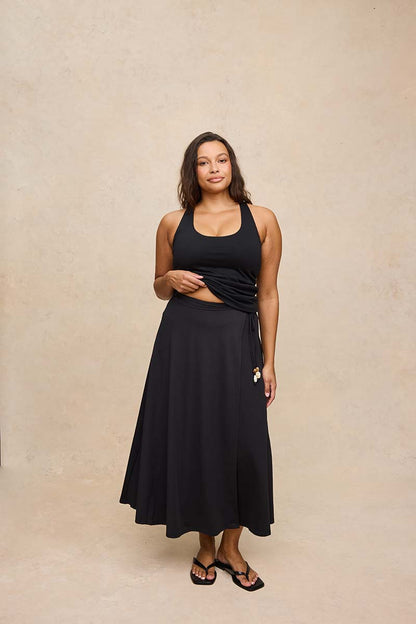 A woman modeling a black two-piece set consisting of a halter crop top and a matching high-waisted midi skirt, standing against a textured beige studio backdrop.