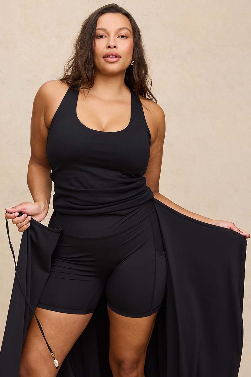 A woman modeling a black sleeveless top holding open a long black draped skirt layer behind her legs, with matching black attached shorts against a warm beige studio backdrop.