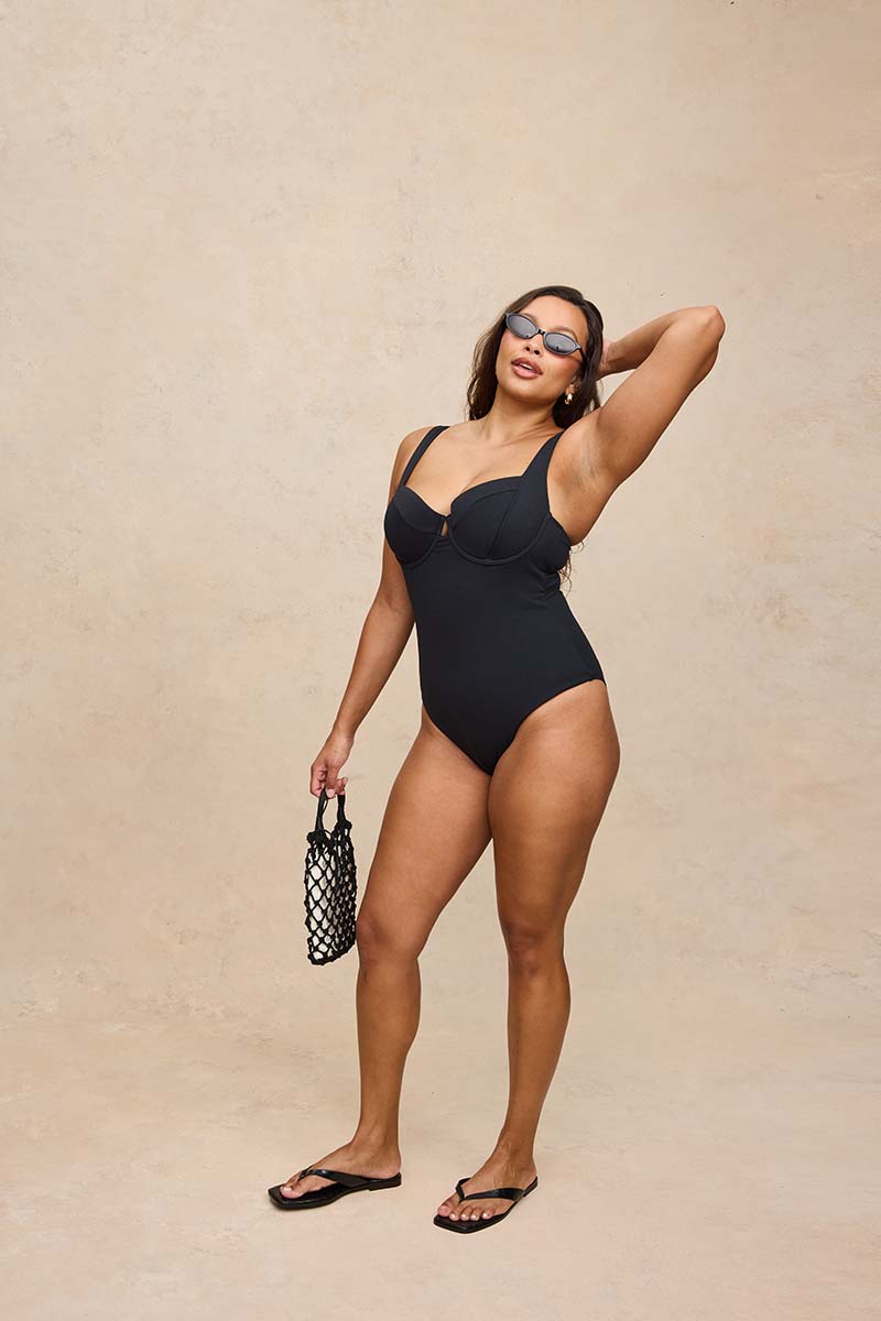 A woman modeling a black one-piece swimsuit with a sweetheart neckline and shoulder cutouts, standing against a textured beige studio backdrop and holding a small black-and-white patterned handbag.