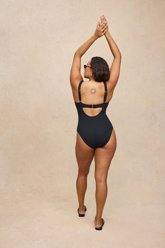 A woman wearing a black one-piece swimsuit is shown from behind, standing against a textured beige studio backdrop with her arms raised overhead.