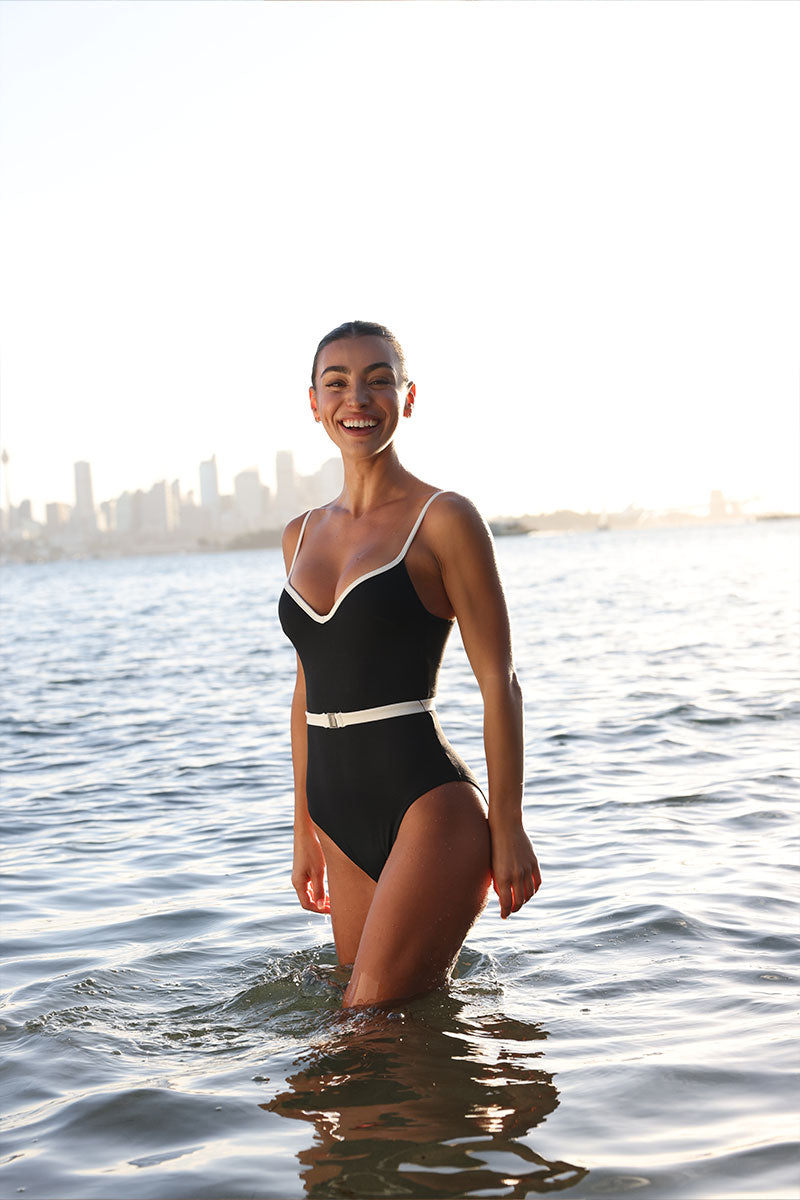 Female wearing a Fearless Swimwear supportive, 360 degree compression black one piece swimsuit with white trims and belt. She stands in the ocean with a city skyline behind her.