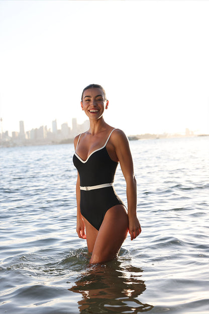 Female wearing a Fearless Swimwear supportive, 360 degree compression black one piece swimsuit with white trims and belt. She stands in the ocean with a city skyline behind her.