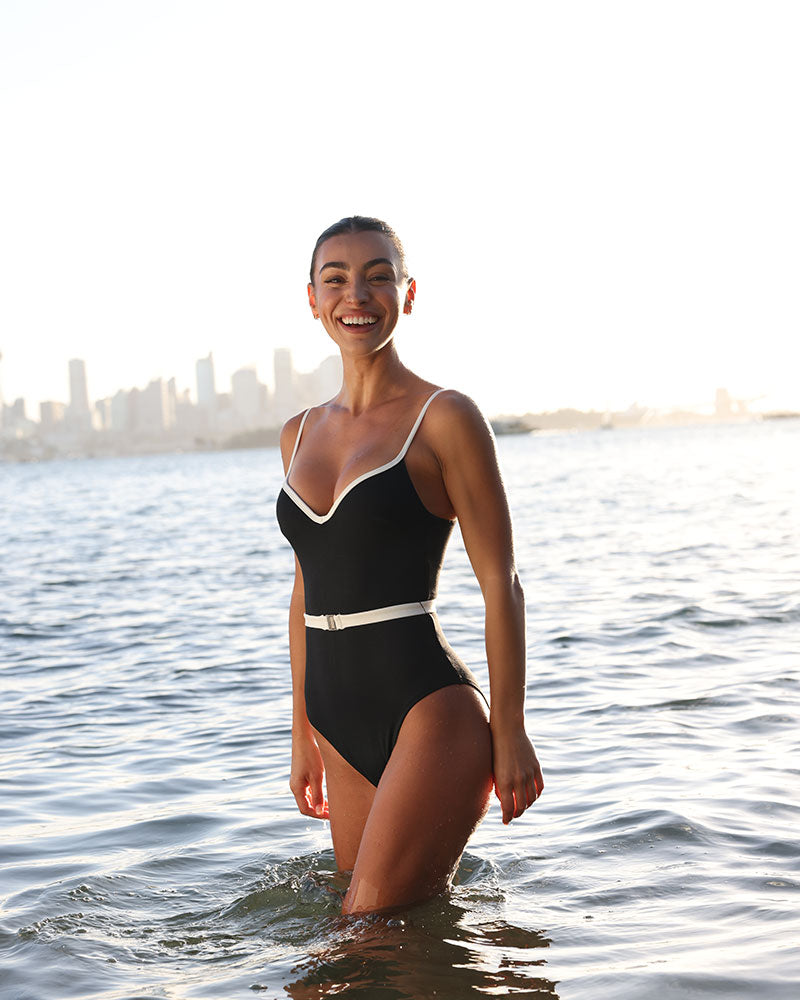Female wearing a Fearless Swimwear supportive, 360 degree compression black one piece swimsuit with white trims and belt. She stands in the ocean with a city skyline behind her.