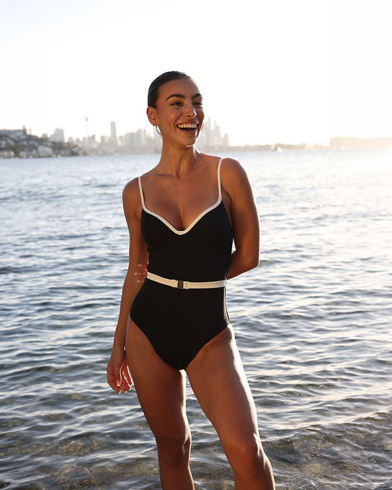 Woman in a black swimsuit standing by water with a city skyline in the background
