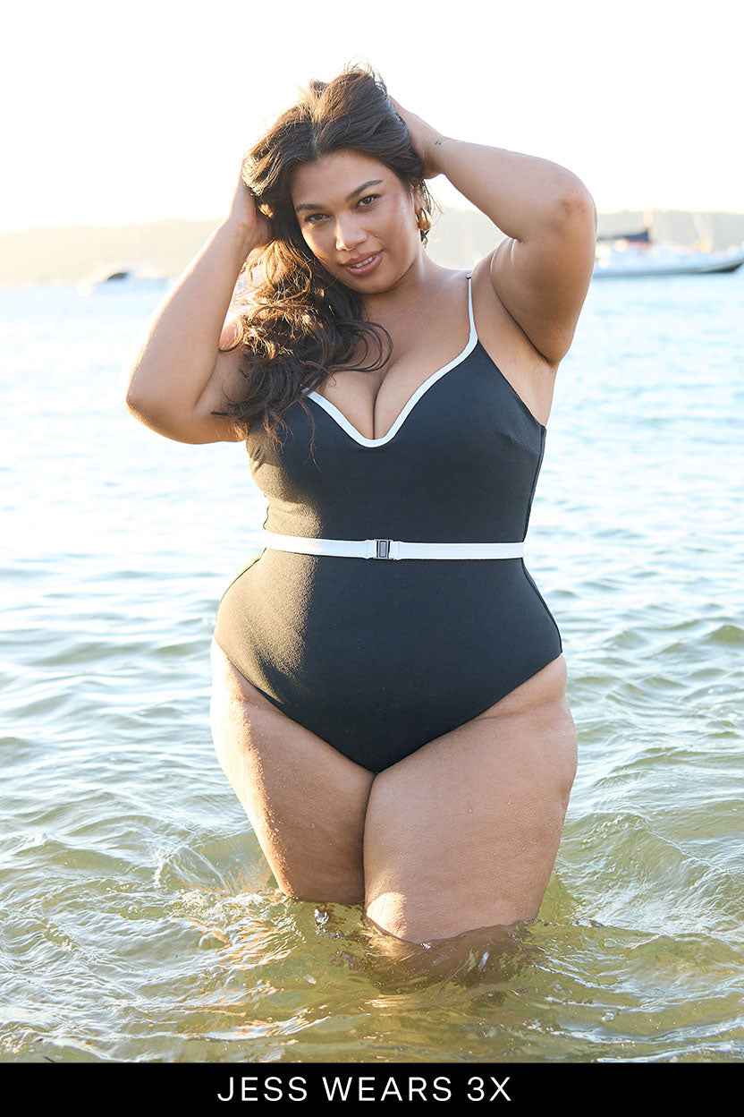 A woman with long dark hair smiles in shallow water wearing the Fearless au Grace High Rise One Piece, a black swimsuit with white trim, a belt, and moulded cups for support. Text at the bottom reads: JESS WEARS 3X.