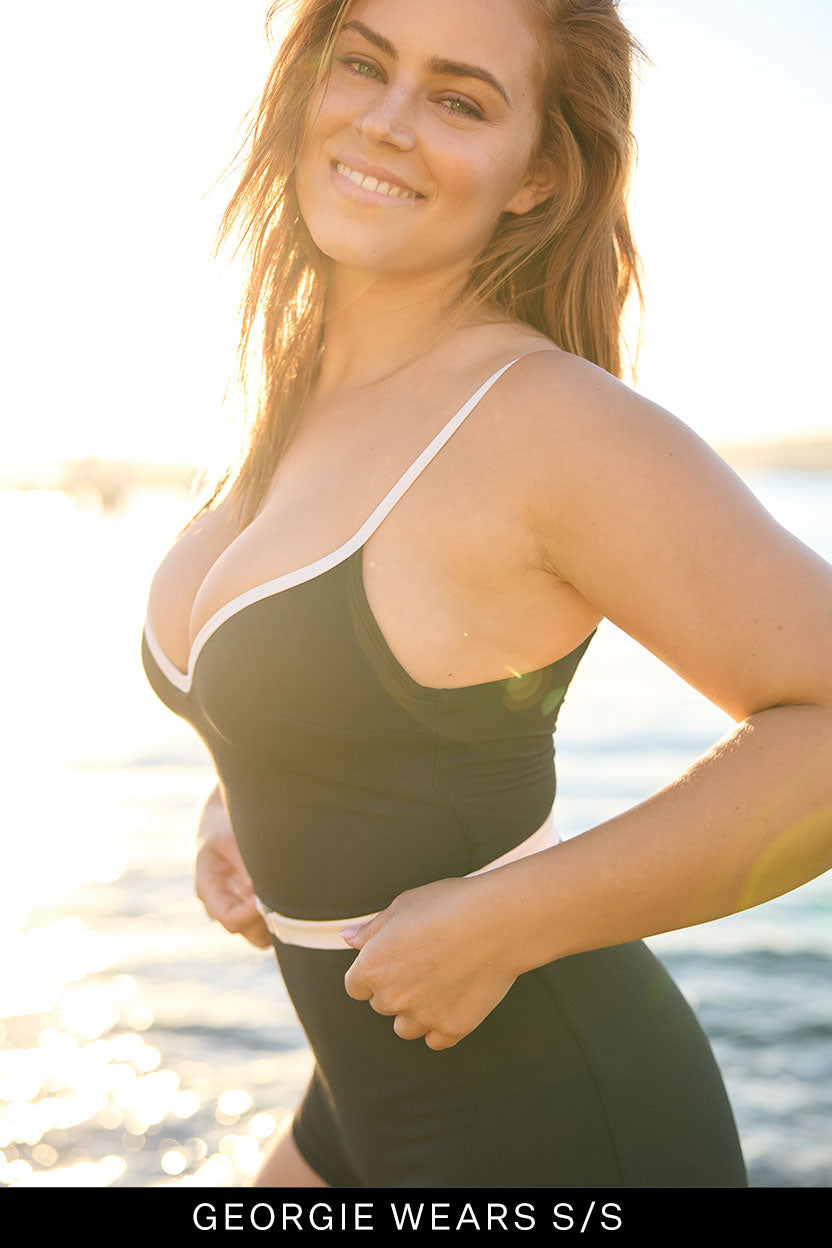 A woman with long brown hair smiles by the water at sunset, wearing the Grace Boyleg One Piece by Fearless au. Text below reads, GEORGIE WEARS S/S.