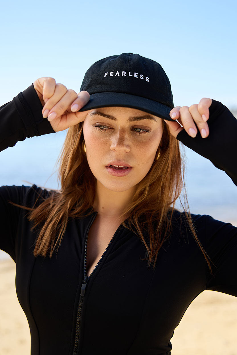 A woman outdoors in a black zip-up top holds the brim of her Fearless au Fearless Cap for sun protection. The background features blurred sand and blue sky.