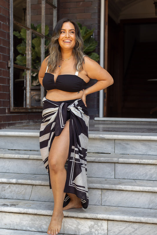 A woman in the Fearless au Geo Print Sarong and a black bikini top smiles as she walks barefoot down marble steps, surrounded by brick walls and lush green plants.