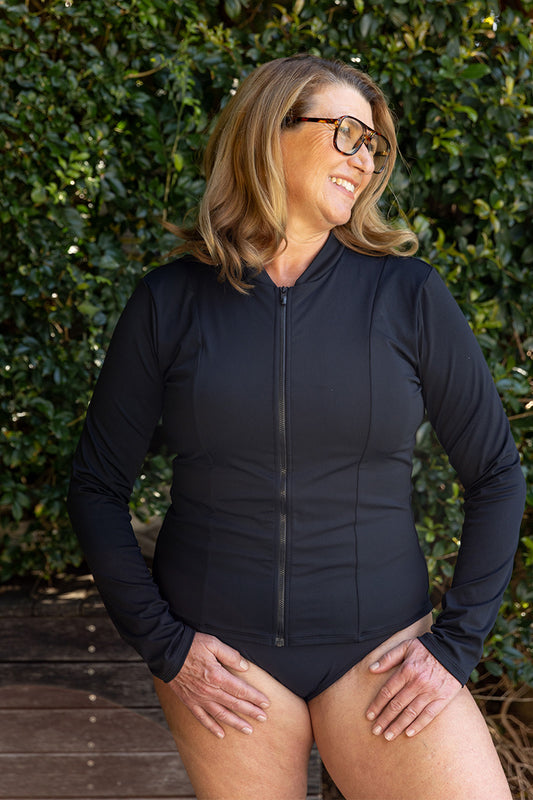A woman with shoulder-length light brown hair and glasses smiles outdoors, wearing the Fearless au Long Rashie with UPF 50+ sun protection and black bikini bottoms, surrounded by green foliage.