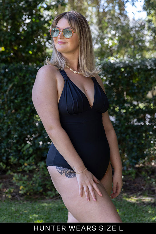 A woman with shoulder-length blonde hair, sunglasses, and a necklace stands outdoors in front of green bushes wearing the Fearless au All Day One Piece - Standard swimsuit in black. Text at the bottom reads: HUNTER WEARS SIZE L.
