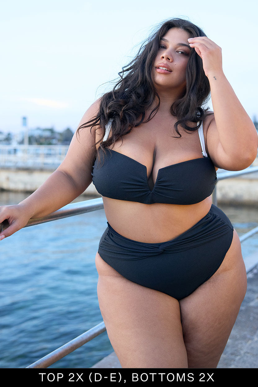 A woman with long dark hair poses by a waterfront wearing the Fearless au Icon Bikini Top in black with white straps, made from recycled crinkle fabric. Text reads: "Top 2X (D-E), Bottoms 2X." Rails and water appear in the background.