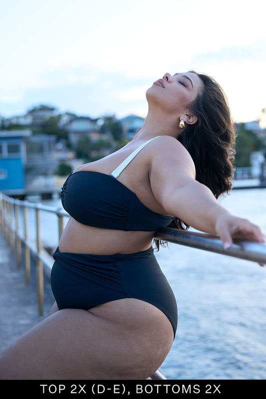A woman poses confidently on a waterfront boardwalk in the Fearless au Icon Bikini Top, black with white straps, made from recycled crinkle fabric. Water and blurred buildings are behind her. Text below reads, TOP 2X (D-E), BOTTOMS 2X.