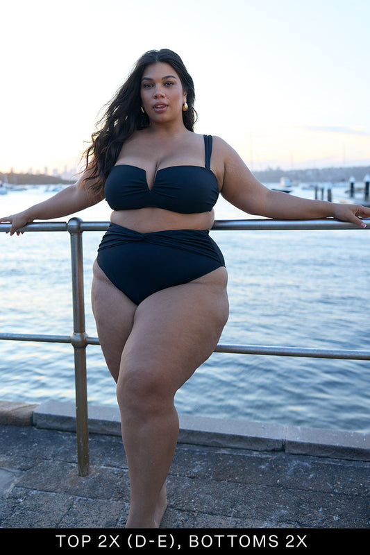 A woman in the Fearless au Icon Bikini Top in black poses by a waterfront at sunset, holding a railing. Text at the bottom reads, TOP 2X (D-E), BOTTOMS 2X. The background features water, boats, and a city skyline.