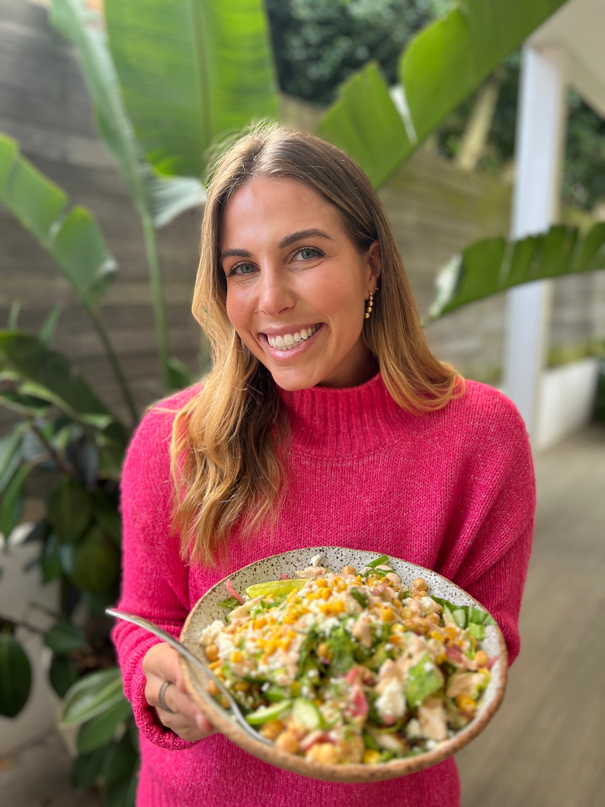 Blonde haired woman wears a pink jumper holding a salad bowl with food in it.