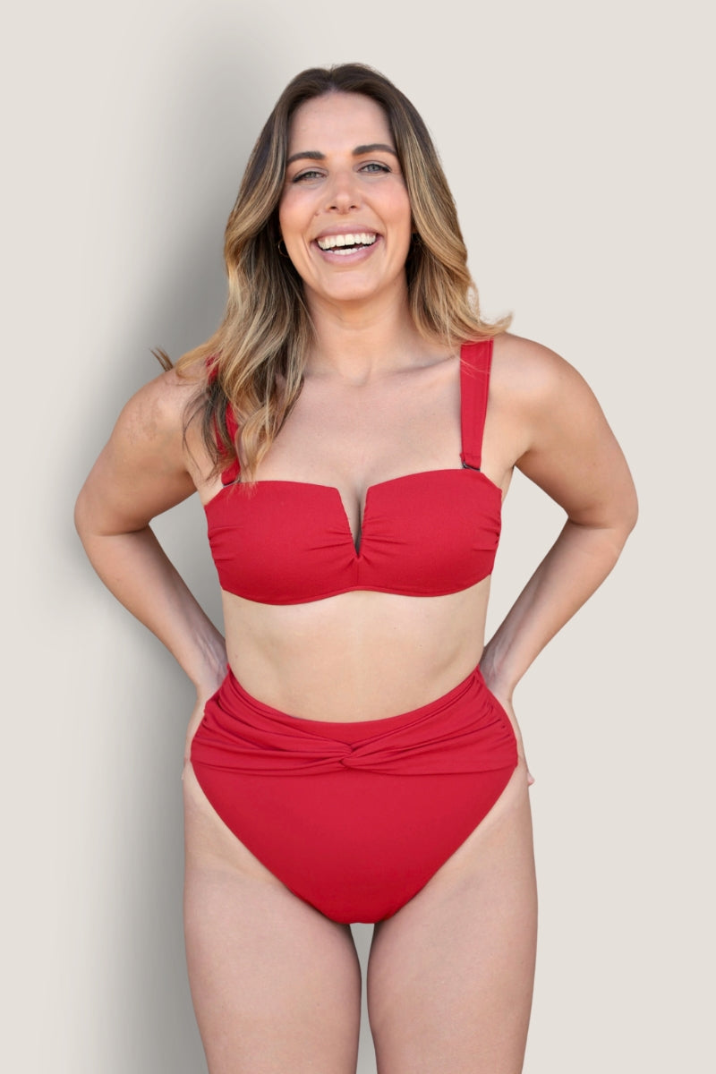 A smiling woman with long, wavy brown hair wears the Fearless au Icon Bikini Top in red, featuring wide straps and a V-neckline, made from recycled fabric, standing against a plain light background.
