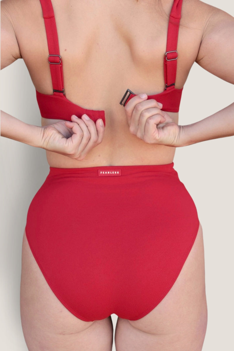 A person seen from the back adjusts the strap of a matching red Icon Bikini Top by Fearless au, paired with a red high-waisted bikini bottom made from recycled fabric.